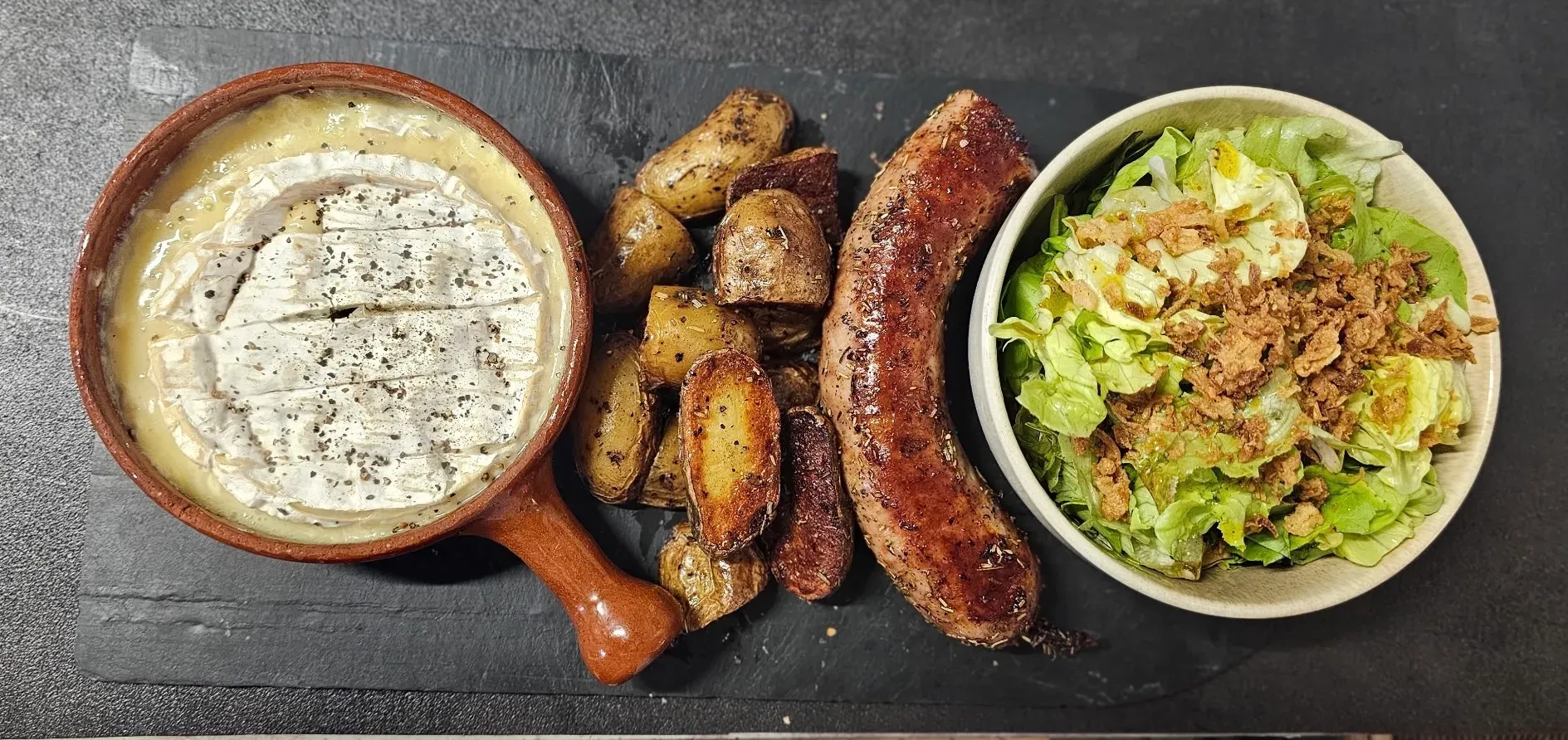 Camembert rôti dans un poêlon en terre, saucisse grillée, pommes de terre grenailles et salade verte, vus de dessus