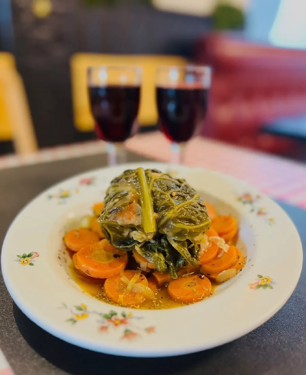Chou farci posé sur des rondelles de carottes dans une assiette fleurie, avec deux verres de vin rouge au fond
