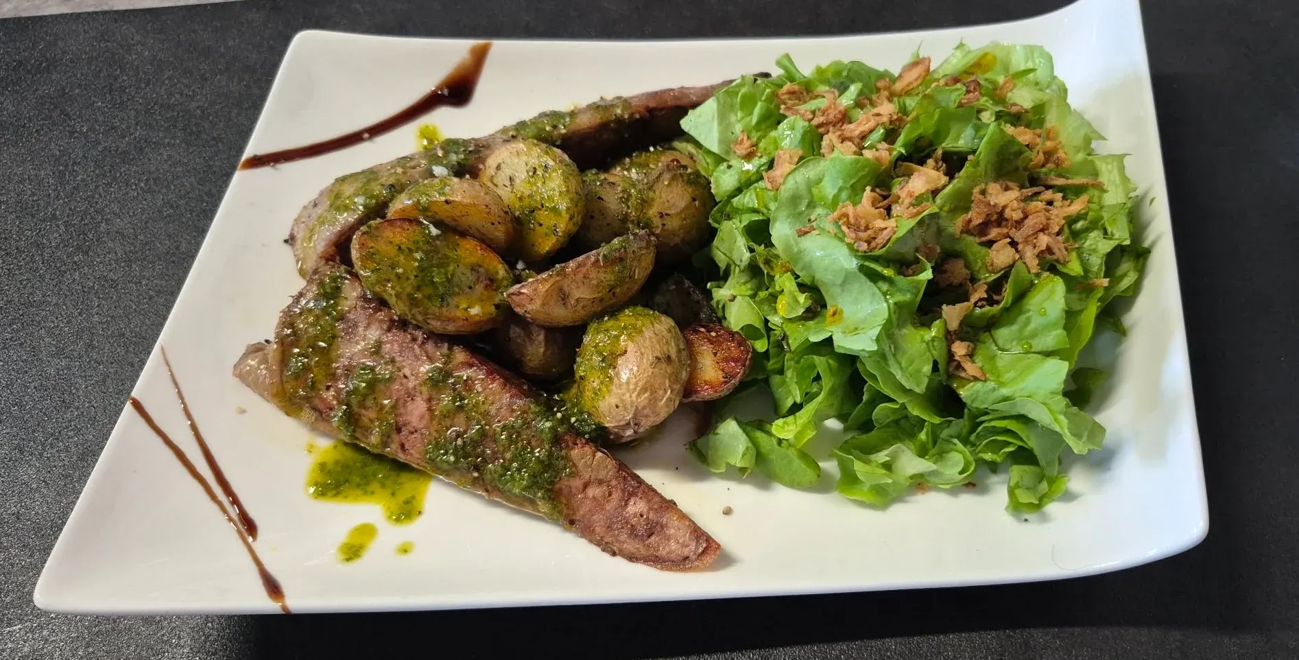 Tranches de cou farci grillé avec huile d’herbes, pommes de terre grenailles et salade verte aux oignons frits, sur une assiette blanche