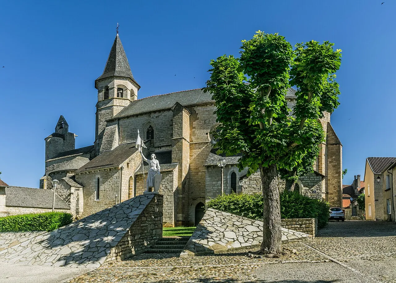 Église du Saint-Sépulcre de Villeneuve-d’Aveyron, en pierre, avec son clocher et une statue blanche au premier plan