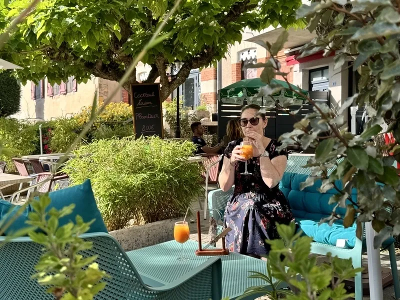 Terrasse pavée ombragée par des arbres, avec fauteuils turquoise, tables et une cliente qui boit un cocktail orange